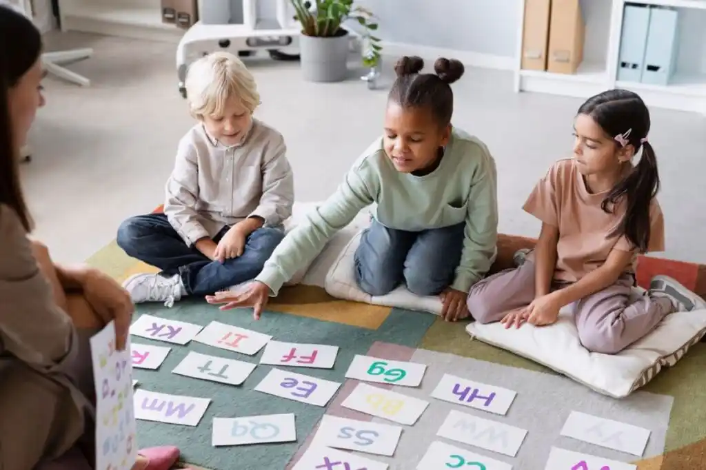 Children engaged in a learning activity, sitting on cushions with alphabet cards spread out before them.