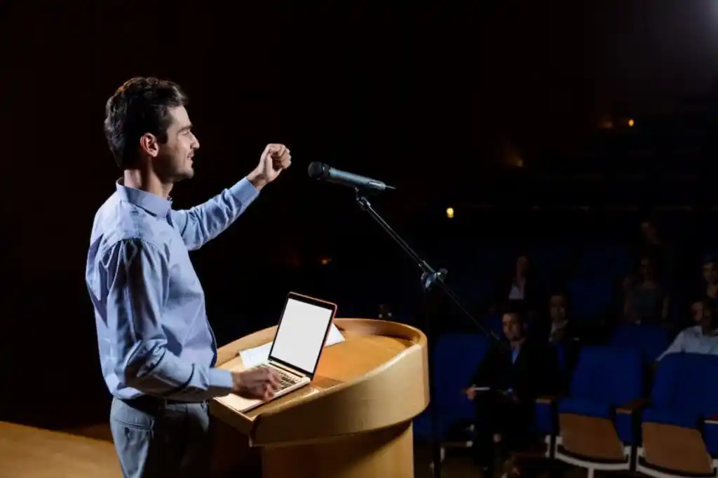 A speaker passionately at a podium, with a laptop and microphone, addressing an audience in a darkened auditorium.