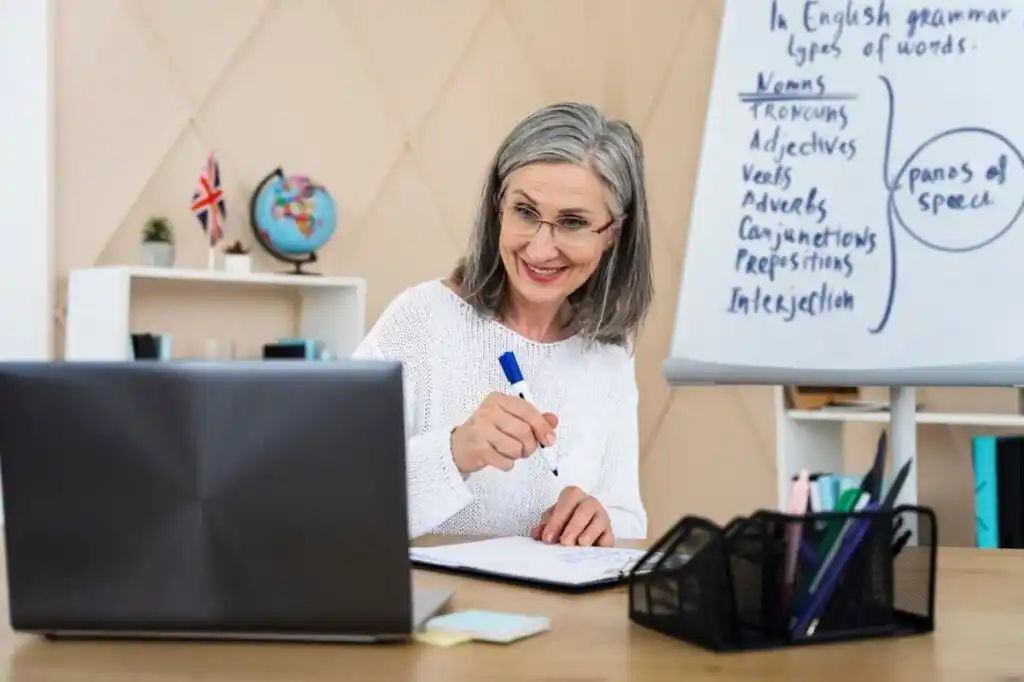 A woman with gray hair writes in a notebook at a desk, with a laptop and a whiteboard