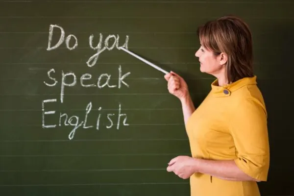 A teacher in a yellow dress points at a chalkboard displaying the phrase "Do you speak English?" in white chalk.