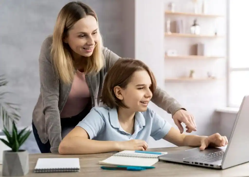 A woman assists a child with a laptop at a desk, surrounded by notebooks and plants, in a bright, modern room.