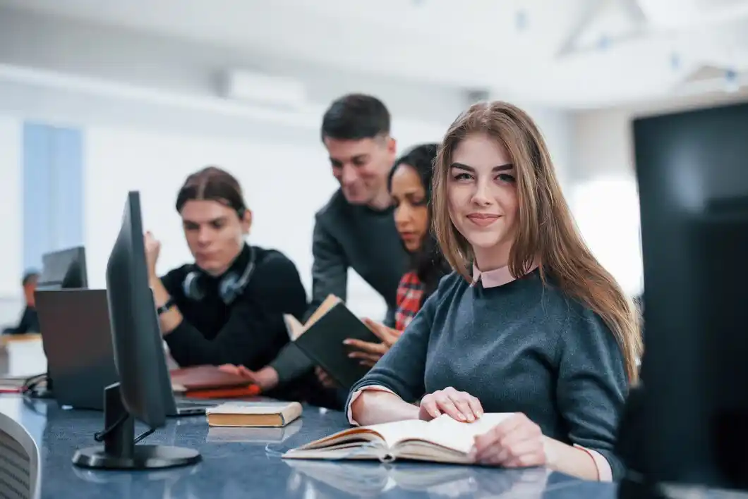 A group of students collaborates in a modern classroom, using computers and books on a sleek table.