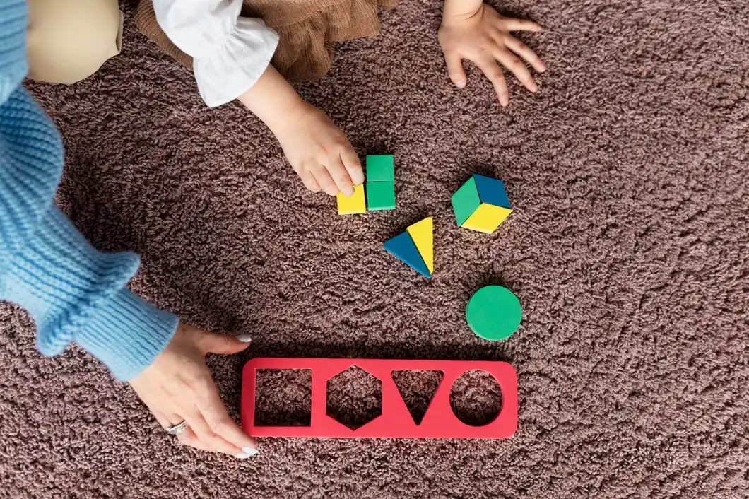 A child's hand reaches for colorful geometric blocks next to a red shape sorter on a soft, brown carpet.