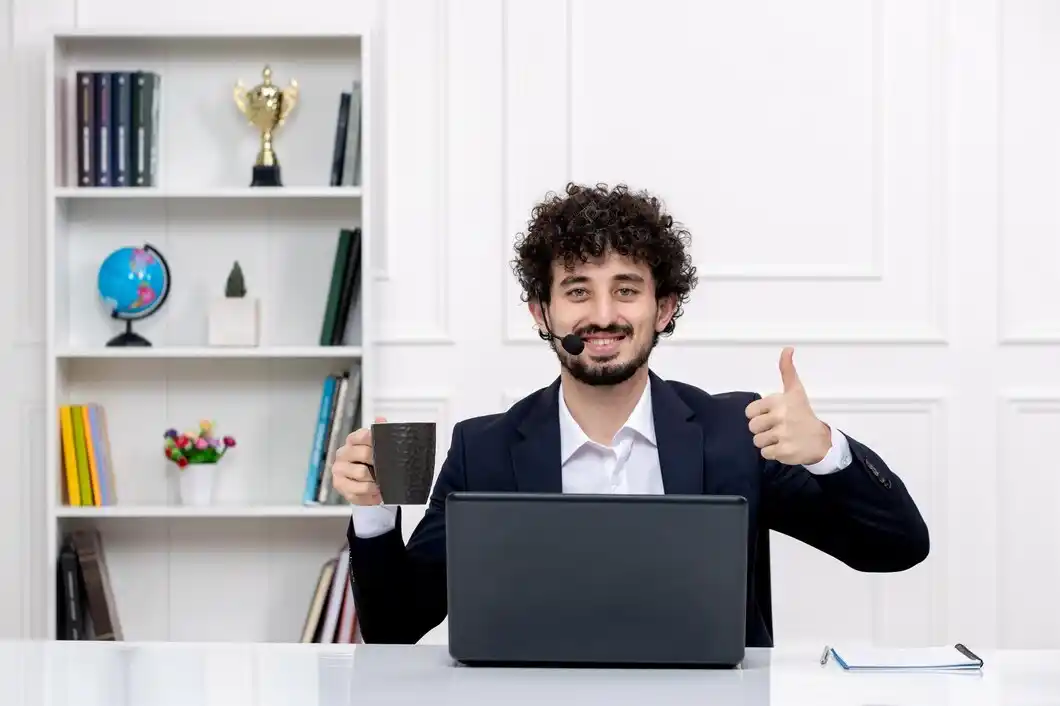 A man in a suit sits at a desk with a laptop, holding a coffee cup and giving a thumbs-up.