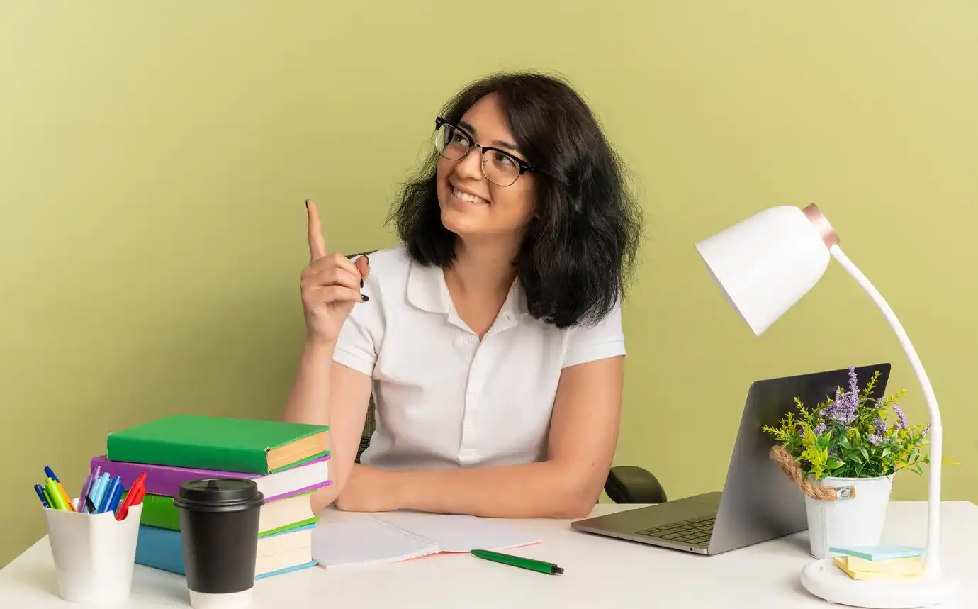 A woman sits at a desk with colorful books, a laptop, and stationery, raising her finger as if making a point.