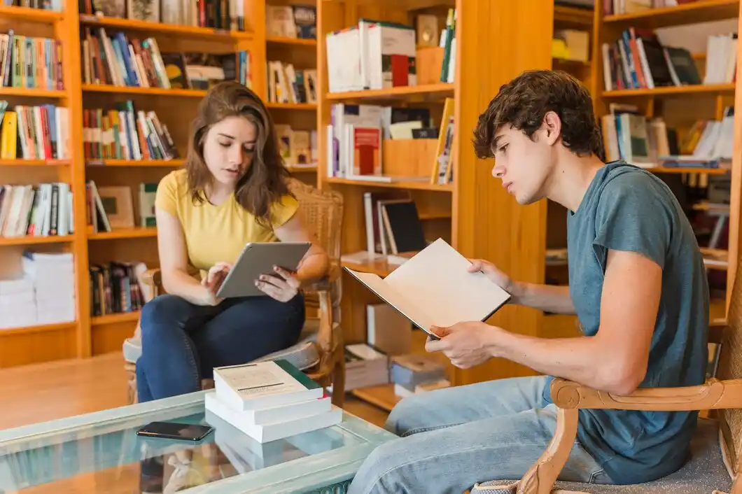 A woman and a man reads a book in a cozy library filled with bookshelves and a glass table.