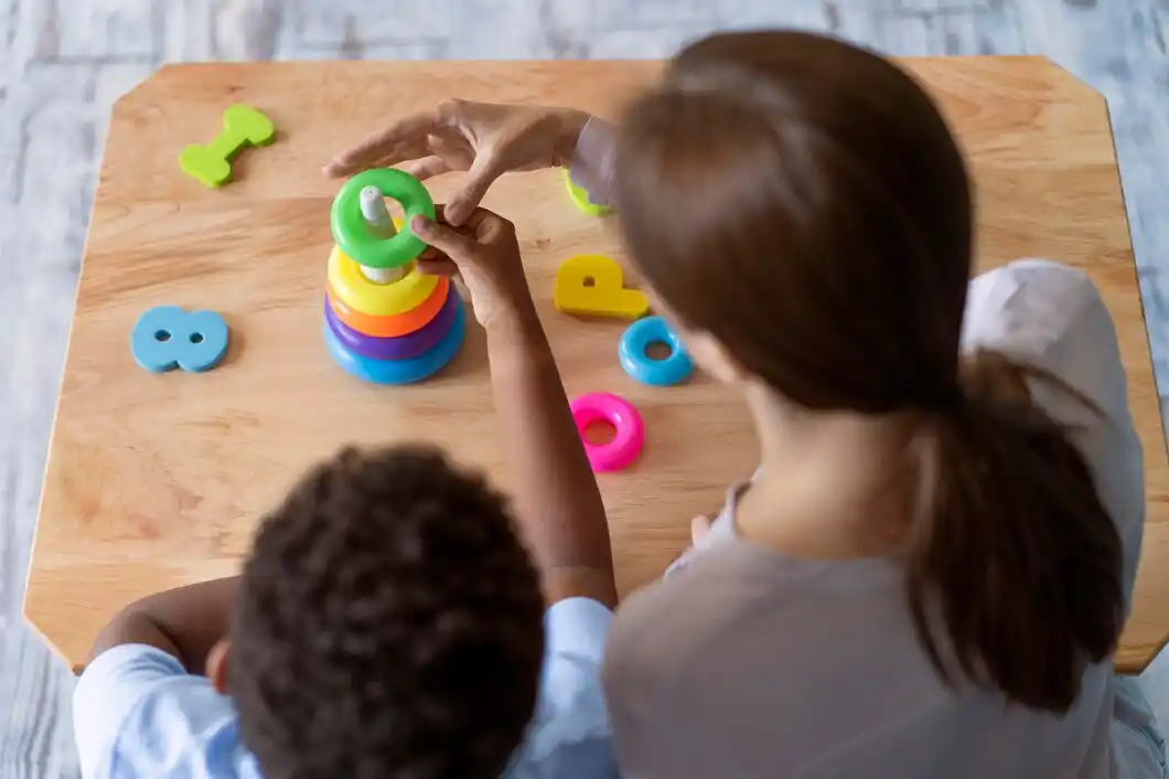 A child play, stacking colorful rings on table, surrounded by additional playful shapes and letters.