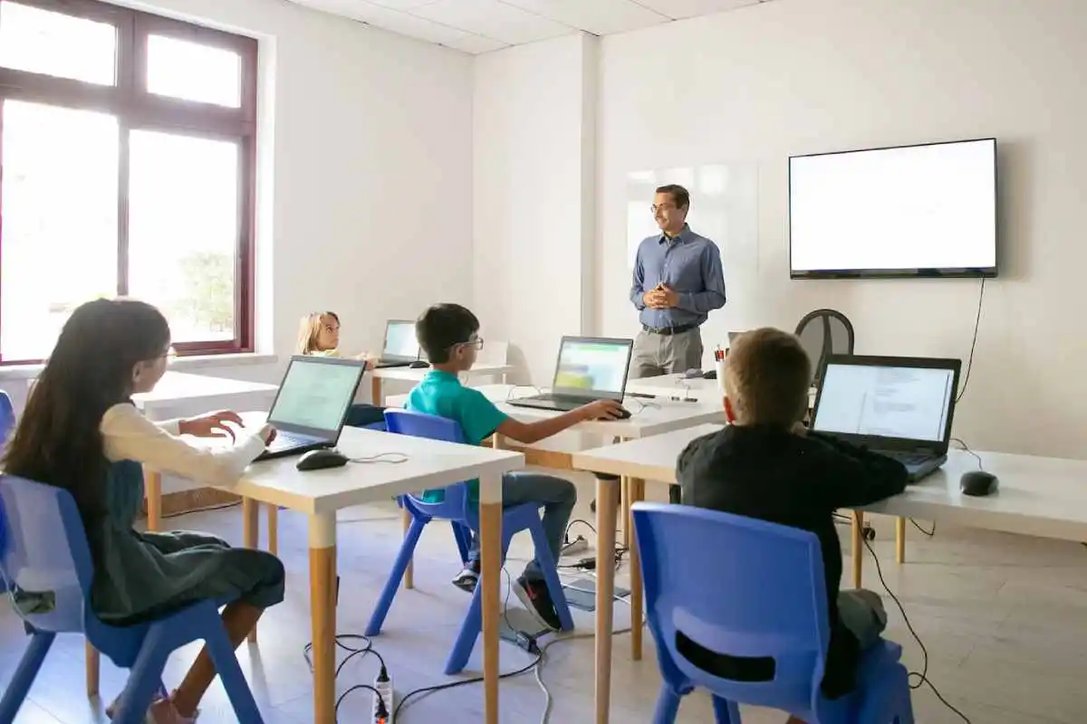 teacher stands before students seated at tables, engaged with laptops