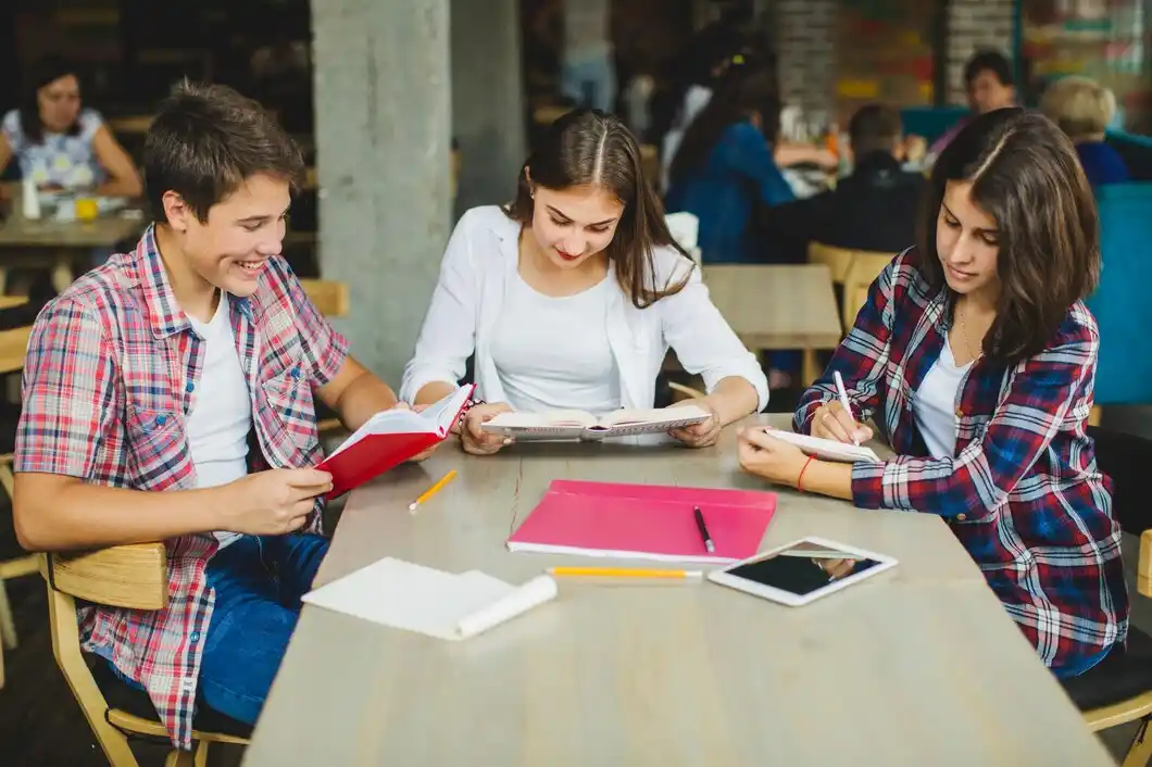 Three students are studying together at a wooden table, surrounded by books and stationery in a busy café setting.