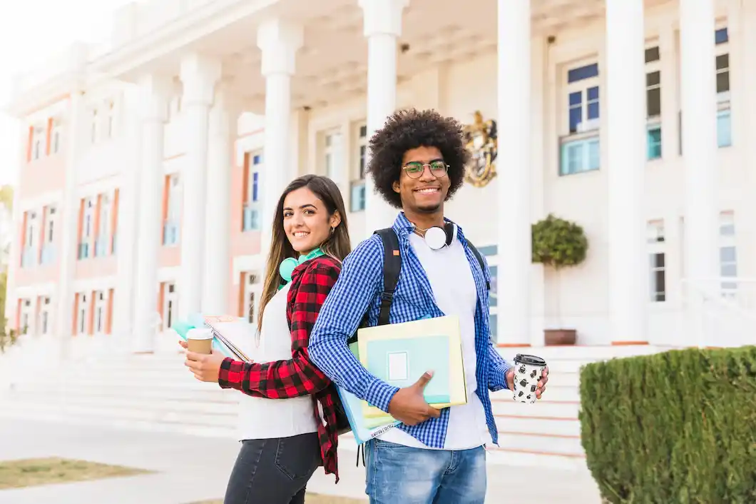 Two students stand in front of a university building, holding books and coffee cups, dressed in casual attire.