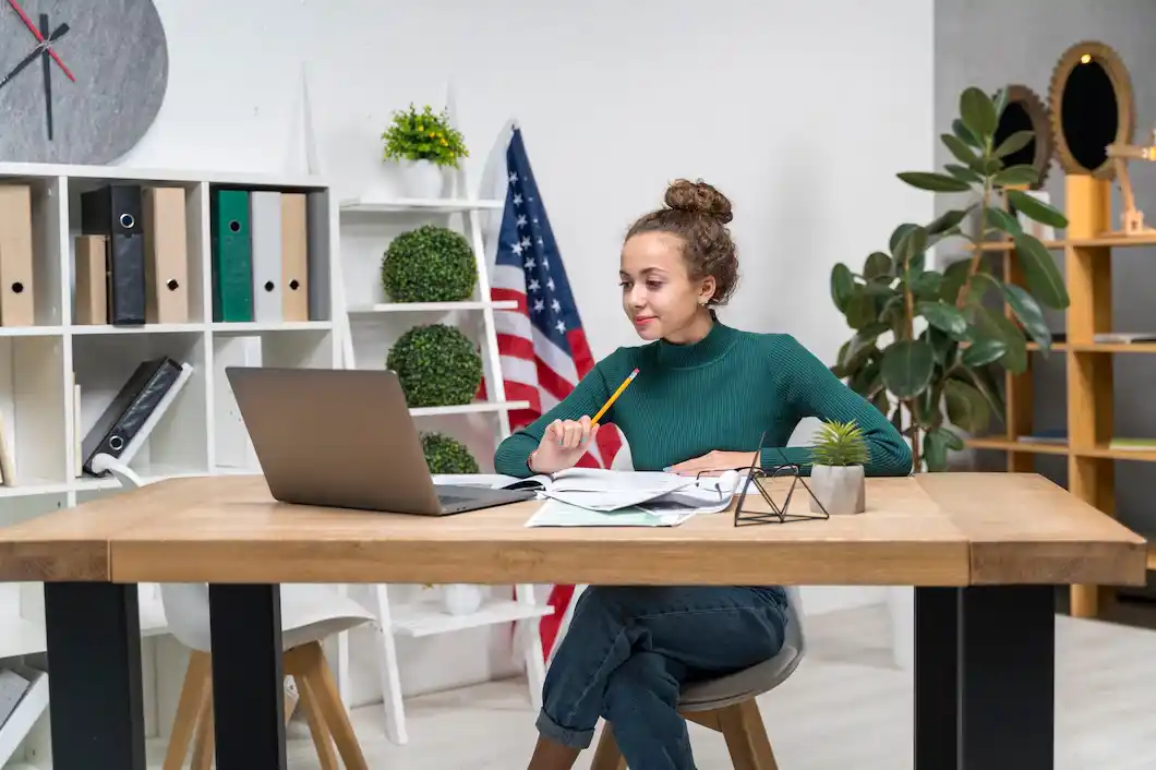 A person in a green sweater sits at a wooden desk, studying with a laptop,