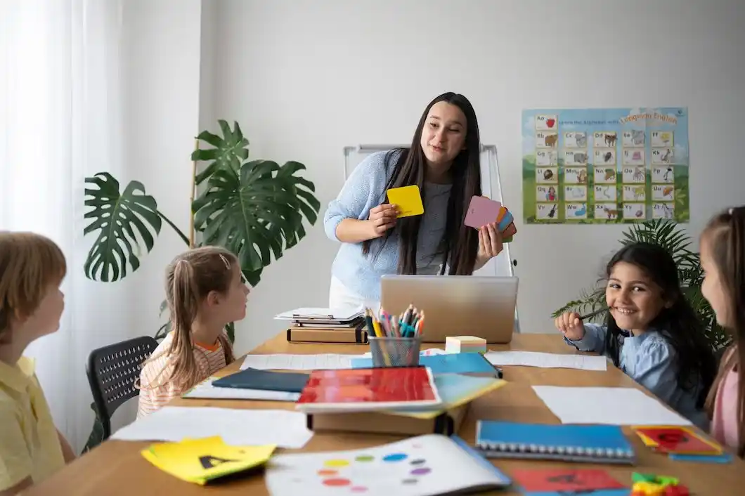 A teacher holds colorful cards during a classroom activity, while children attentively engage at their desks surrounded by learning materials.