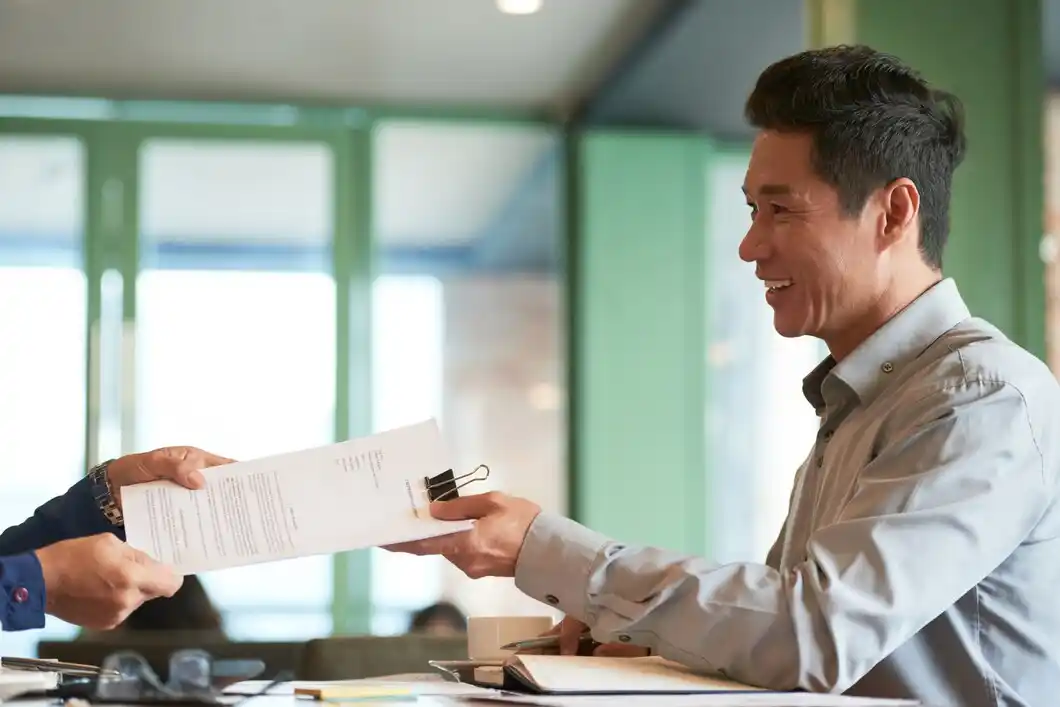 A person in a blue shirt hands over documents to another individual in a light gray shirt at a modern workspace.