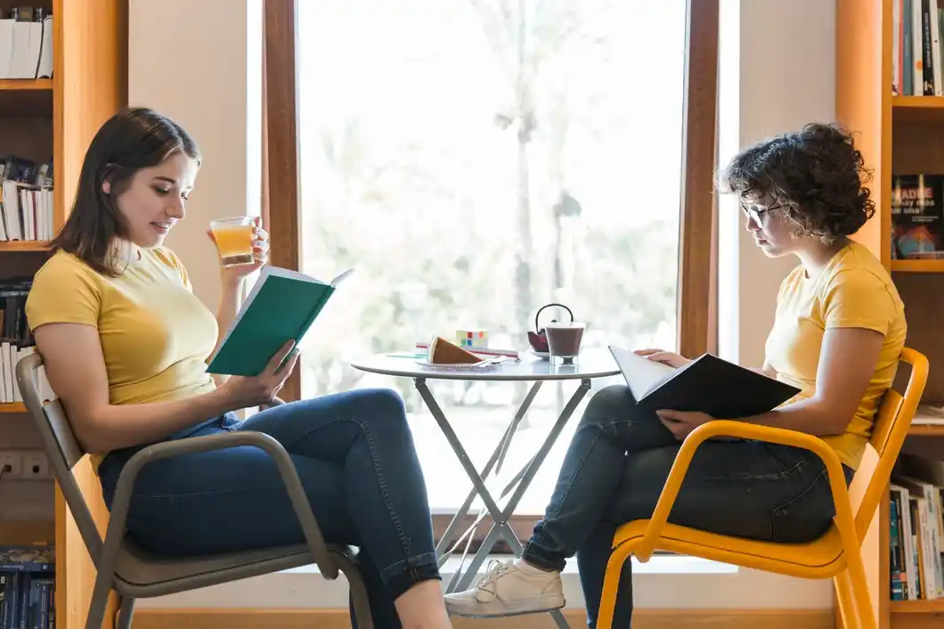Two women reading books and enjoying drinks by a sunlit window with a view of palm trees.