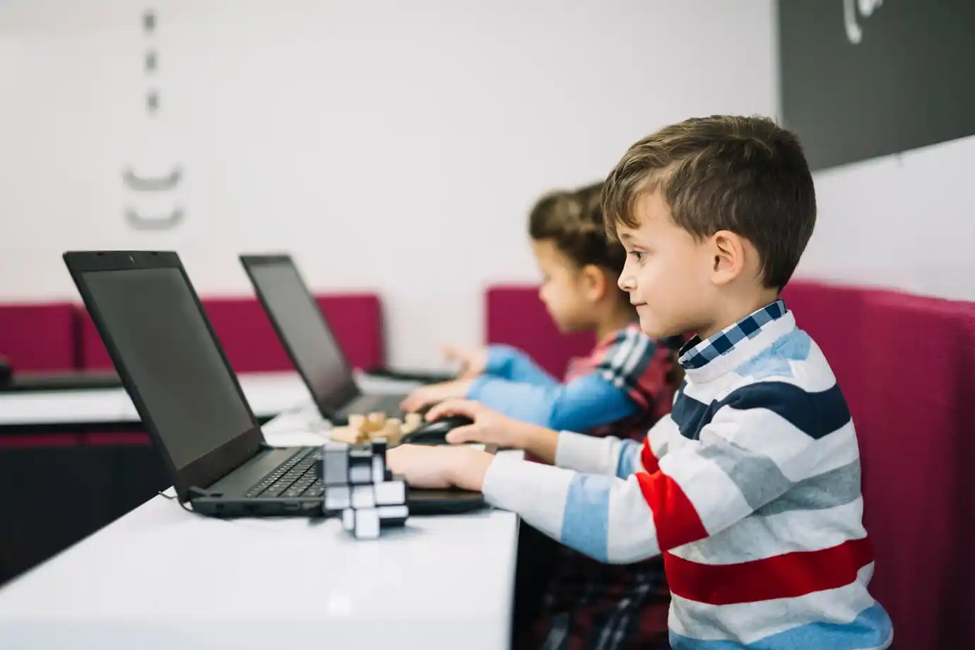 Two children working on laptops at a white table, focused on their tasks